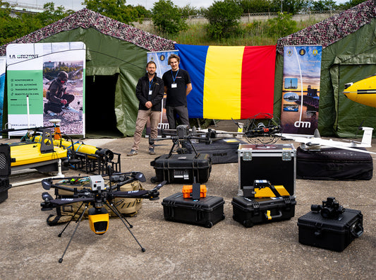 Expoziție de echipamente UAV în România, cu drone industriale, roboți subacvatici și standuri de prezentare La Orizont și Skyline Drones, în fața unui cort militar și a steagului național.